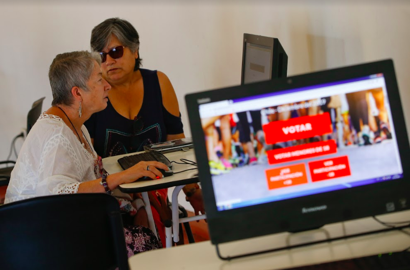 Dos mujeres votando electrónicamente y una pantalla de computador con la plataforma de votación de EVoting
