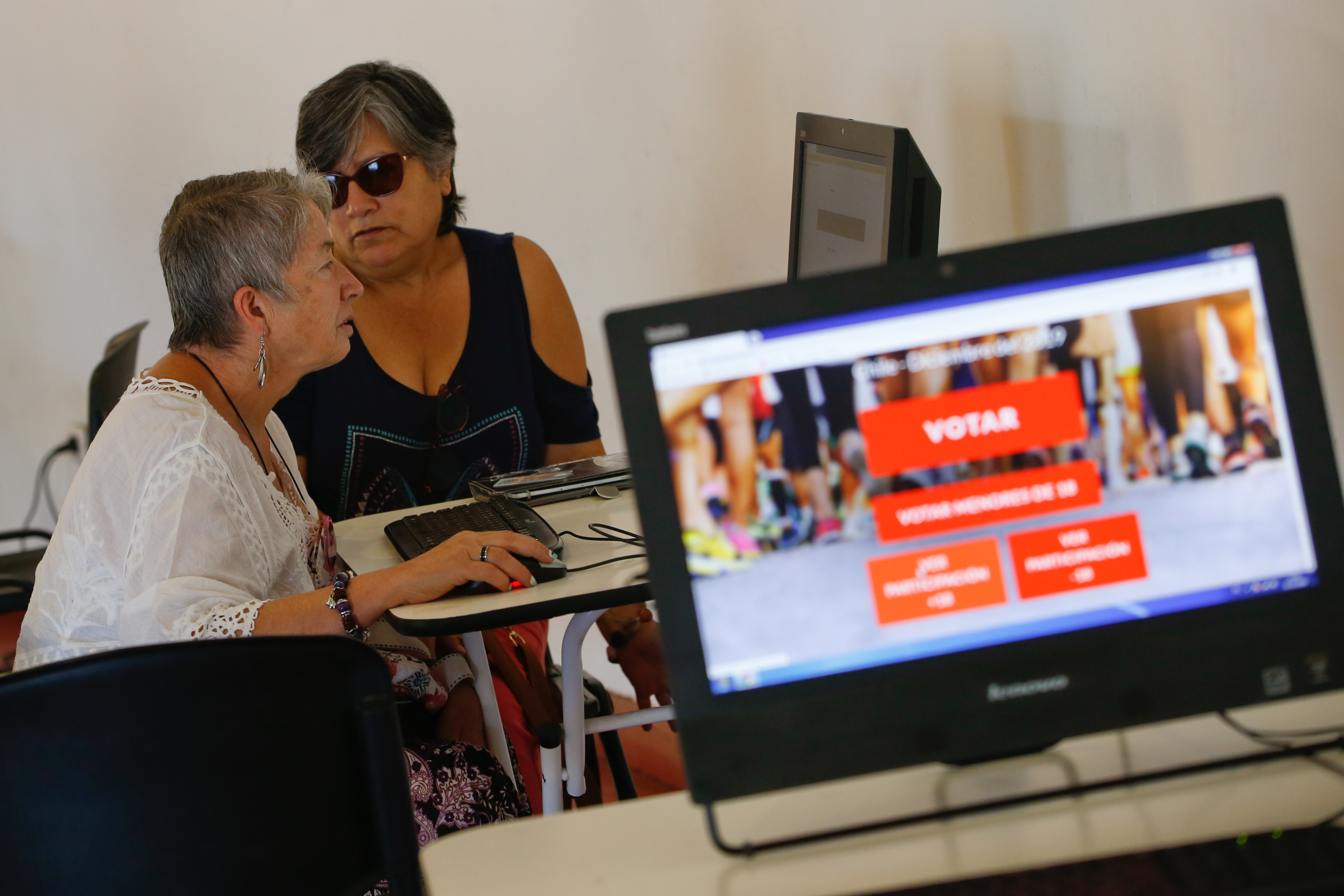 Dos mujeres votando electrónicamente con pantalla de computador que muestra plataforma de EVoting